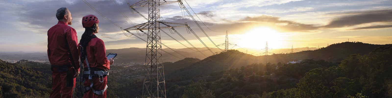 Two workers in red boiler suits watch the sun set over pylons